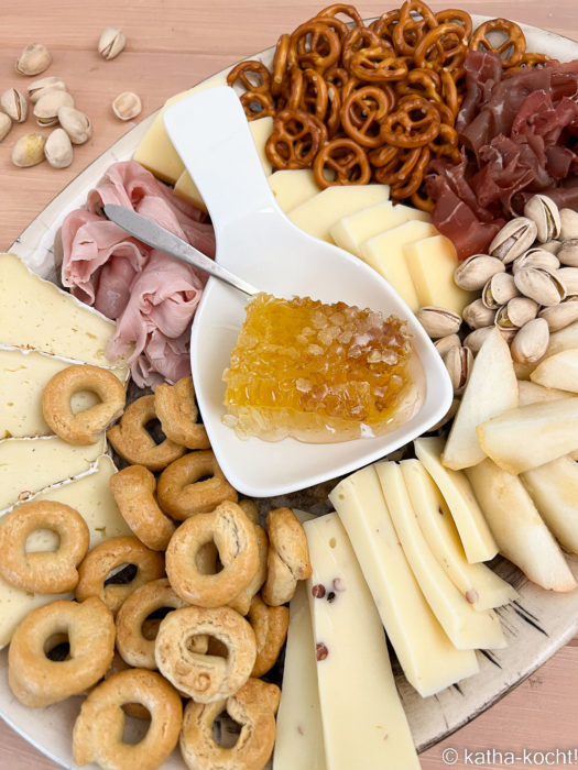 Elegante Charcuterieplatte in hellen Farben mit Käse, Kochschinken, Crackern, und einer Honigwabe in der Mitte der Platte
