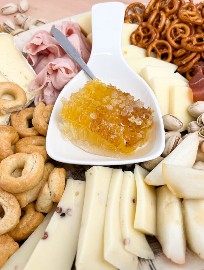 Elegante Charcuterieplatte in hellen Farben mit Käse, Kochschinken, Crackern, und einer Honigwabe in der Mitte der Platte